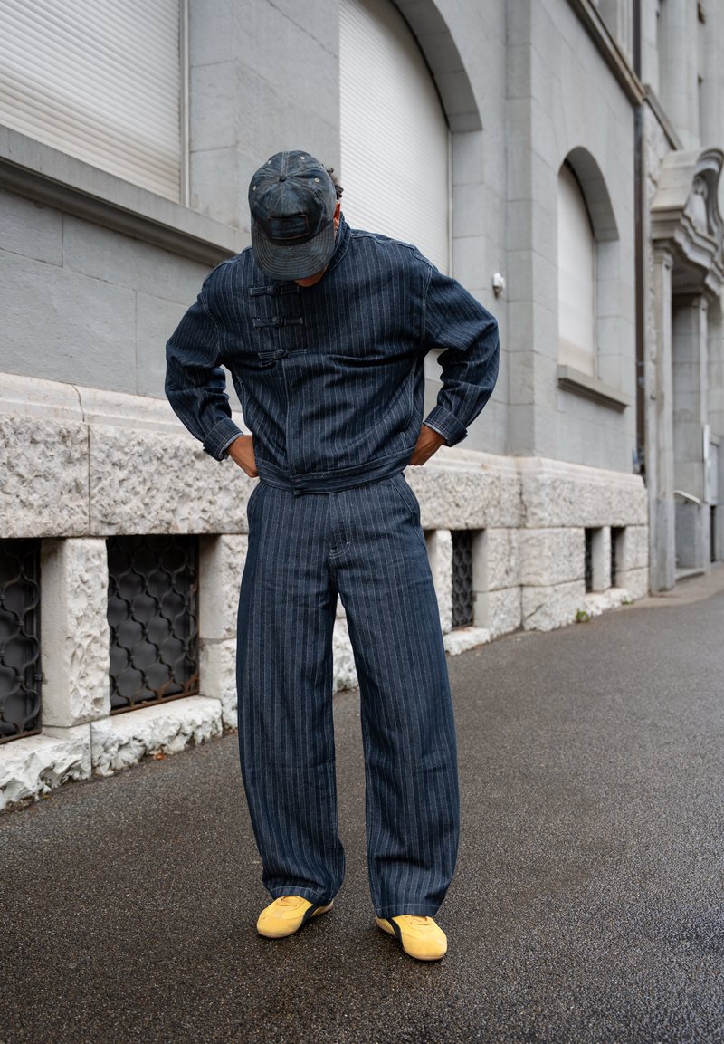 Person wearing dark pinstripe jacket and pants with yellow shoes and a dark cap, standing on a wet street near a stone building.