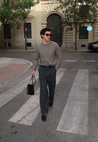 Vīrietis valkā saulesbrilles, pelēku džemperi un tumšas bikses, iet pa pilsētas gājēju pāreju, turēdams melnu soma, fonā redzami koki un ēkas.