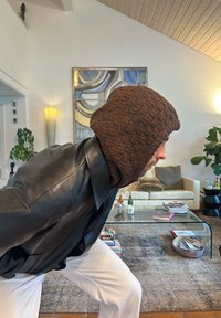 Brązowa, dziergana czapka z wyraźnym wzorem, noszona z czarną skórzaną kurtką. Widoczne białe spodnie. Nowoczesne wnętrze.