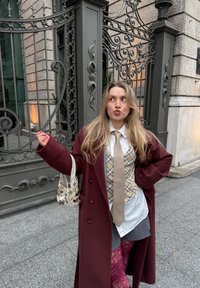 Manteau marron, cravate beige, gilet à carreaux, chemise blanche. Leggings roses texturés et sac à main à perles. Décor extérieur avec une porte décorative.