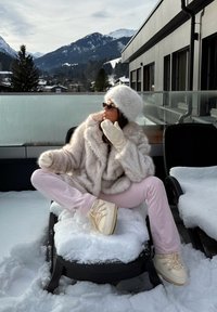 Ženska v belem krznenem plašču, klobuku, rokavicah in rožnatih hlačah sedi na stol, pokrit s snegom, na zasneženem balkonu z gorami v ozadju.