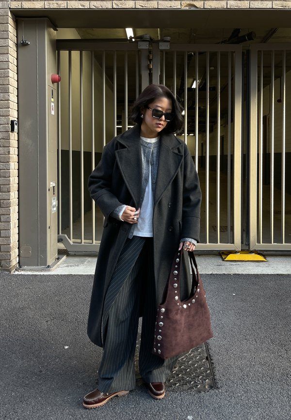 Femme portant un manteau sombre oversize, un cardigan gris, un pantalon à fines rayures, des mocassins, des lunettes de soleil, tenant un sac cabas marron clouté, debout près d'une entrée grillagée.
