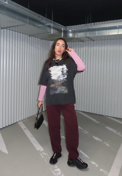 Joven de pie en un estacionamiento, vistiendo una camisa de manga larga a rayas superpuesta con una camiseta gráfica, pantalones burdeos, zapatos negros, y sosteniendo un pequeño bolso negro.