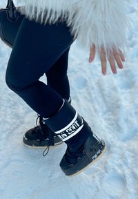 Bottes de neige noires avec une sangle blanche, un design à lacets et des semelles en caoutchouc ; associées à des leggings noirs sur la neige. Vêtement extérieur blanc duveteux partiellement visible.