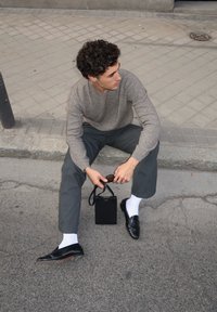 Pelēks garām piedurknēm džemperis, ogļu krāsas svītrainas bikses, melni ādas apavi, balti zeķi; rokās tur mazu melnu somu ar rokturīti.