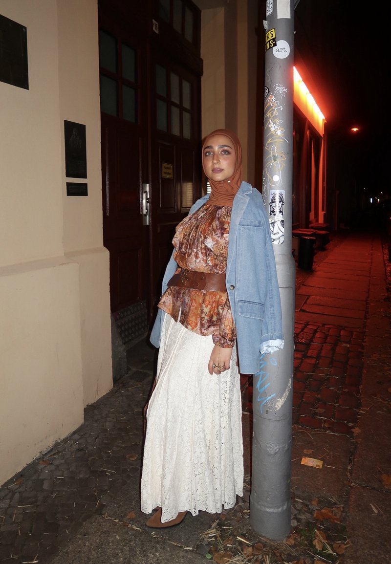 Bluzka typu peplum w kwiatowy wzór, na długiej białej koronkowej spódnicy, zestawiona z dżinsową kurtką, brązowym paskiem i brązowymi szpilkami. Miejskie nocne otoczenie.