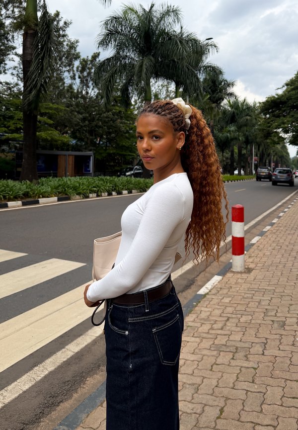 Femme aux longs cheveux bouclés, portant un haut blanc et une jupe foncée, se tient près d'un passage pour piétons dans une rue urbaine bordée de palmiers et de voitures.