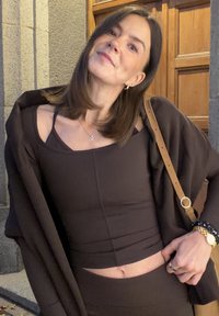Smeđi rebrasti top bez rukava s krojenjem iznad struka, složen ispod labave smeđe vanjske odjeće. Istaknute narukvice i torba za rame s zlatnim akcentom.