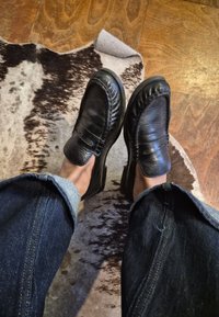 Juodos odinės loafers su austa detale viršuje, padėtos ant medinių grindų, derinamos su tamsaus džinso atloštais džinsais.