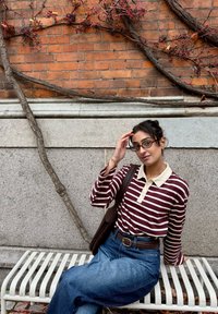 Bordeauxrode en wit gestreepte shirt met lange mouwen en een crèmekleurige kraag, gecombineerd met blauwe high-waisted jeans. Bruine tas en zittend op een witte bank.