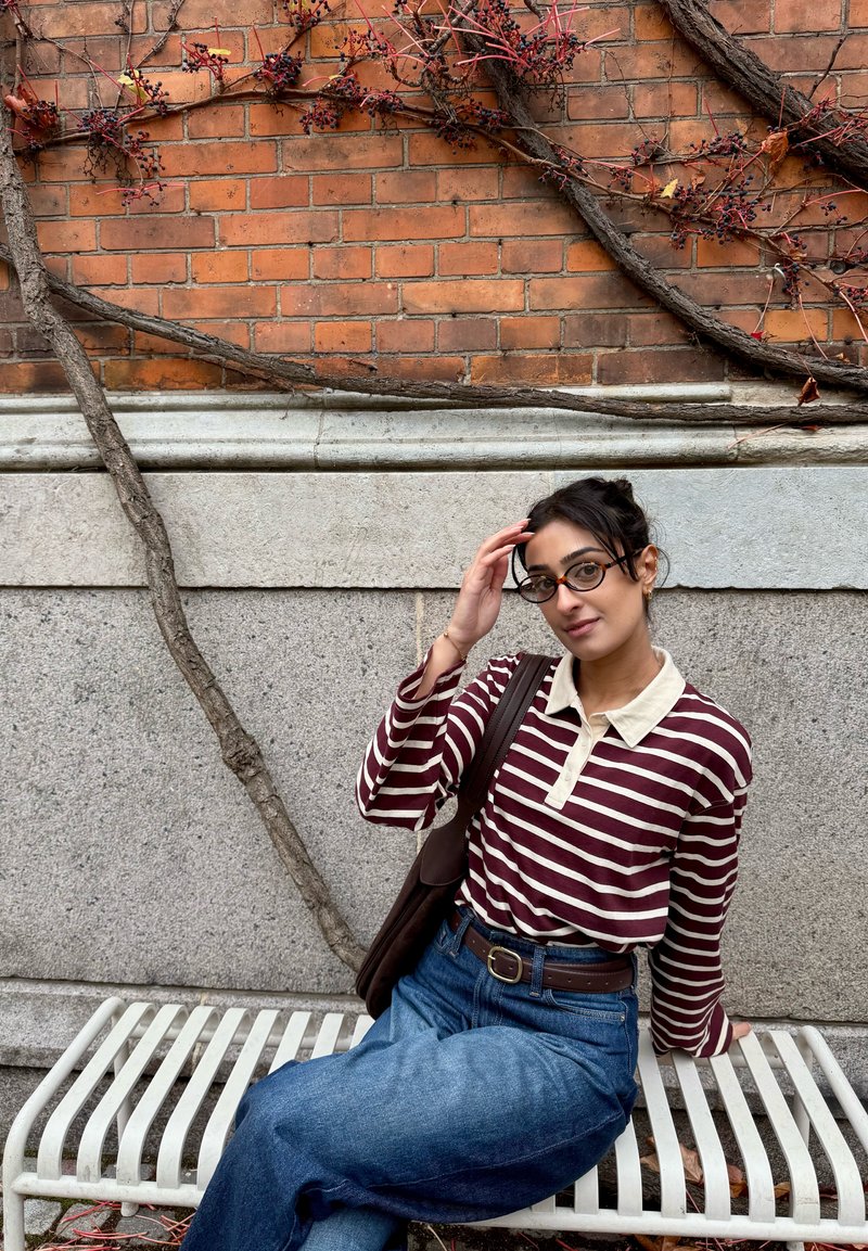 Maglia a maniche lunghe a righe bordeaux e bianche con colletto color crema, abbinata a jeans a vita alta blu. Borsa marrone, seduta su una panchina bianca.