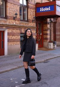 Veste noire en tissu texturé avec des détails frangés, portée sur une robe noire ajustée. Grandes bottes en caoutchouc noires et un petit sac à main noir. Cadre urbain.