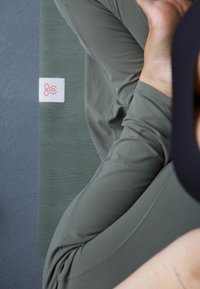 Olíva zöld jóga matrac, texturált felülettel és fehér címkével, amelyen piros grafika található. Rugalmas, illeszkedő anyag egy azonos színben.