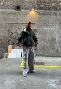 Roheline fleece'i lukuga jakk valge kapuutsiga, hallid taskutega cargo püksid, ruudulised libisevad kingad, väliskeskkonnas graffiti taustal.
