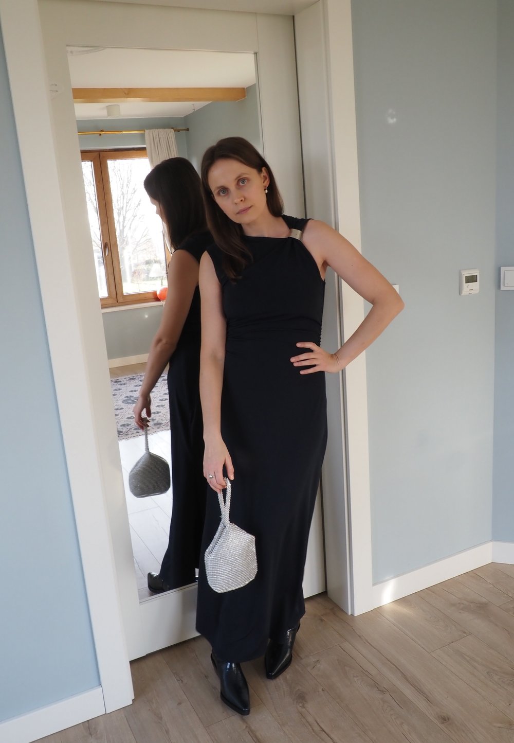 Robe maxi bleu foncé avec une bretelle texturée, associée à un petit sac à main argenté et des bottines noires. Fond en bois et murs clairs.