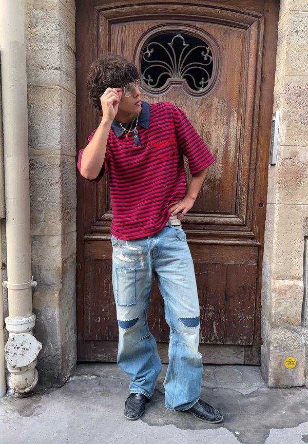 Giovane con occhiali, maglietta a righe rosse e nere e jeans azzurri con toppe, in piedi davanti a una porta di legno con lavorazioni metalliche ornamentali.