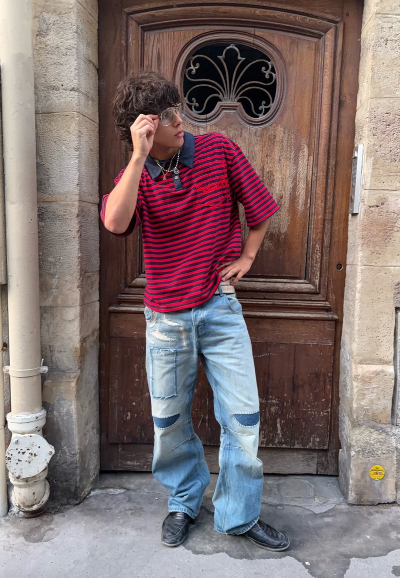 Jong persoon met een bril, een rood-zwart gestreept shirt en lichtblauwe jeans met stukken, staat tegen een houten deur met sierlijk metaalwerk.