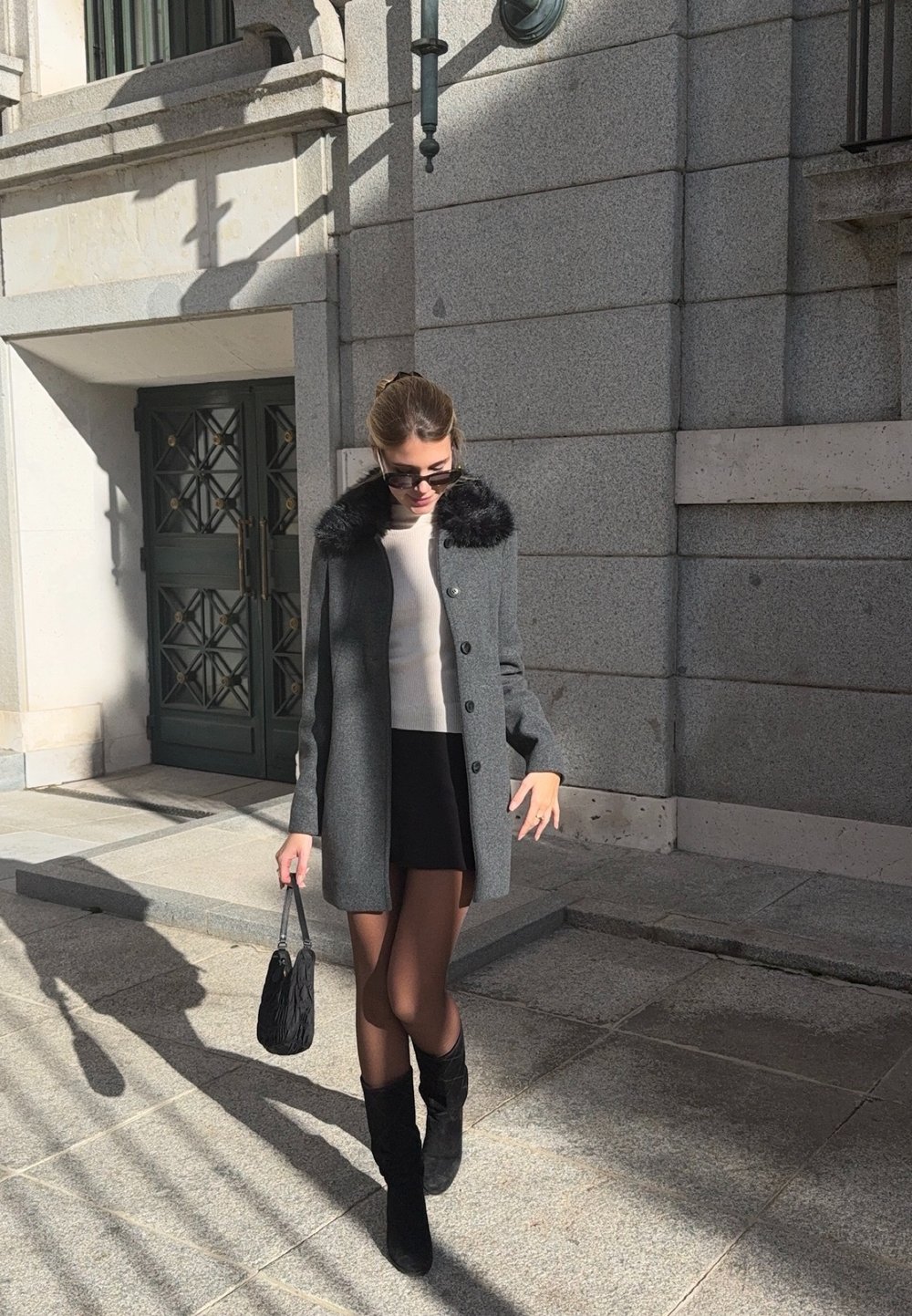 Nő sétál a szabadban, szürke kabátot visel szőrme gallérral, fekete szoknyát, fekete csizmát, napszemüveget, és fekete táskát hord a napfényben.