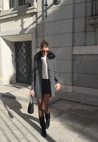 Nő sétál a szabadban, szürke kabátot visel szőrme gallérral, fekete szoknyát, fekete csizmát, napszemüveget, és fekete táskát hord a napfényben.