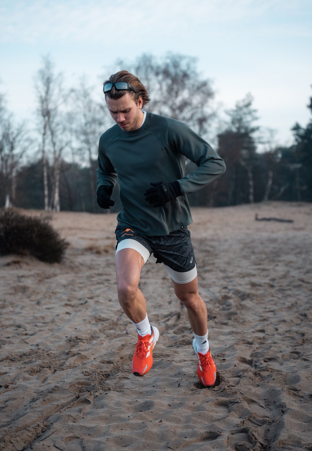 Coureur vêtu d'un haut à manches longues gris et d'un short à motifs, portant des gants noirs et des chaussures orange vif, courant sur un terrain sablonneux.
