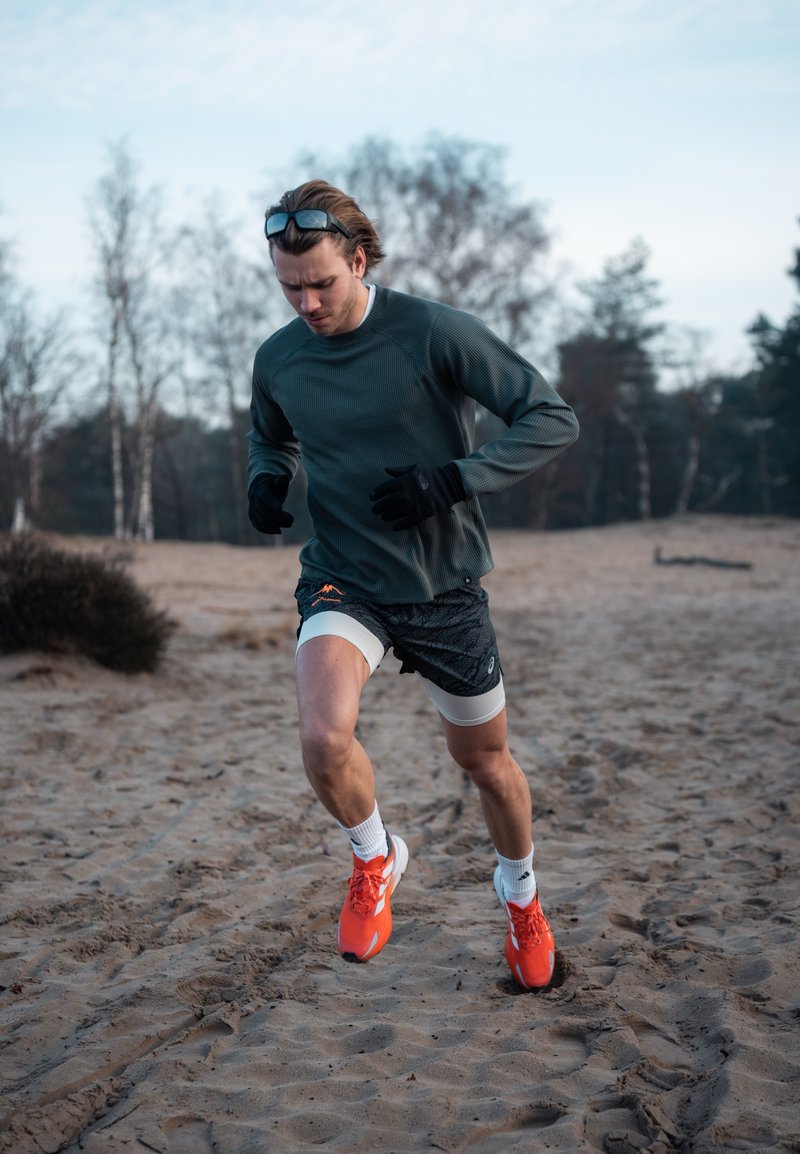 Coureur portant un haut à manches longues gris et un short à motifs, avec des gants noirs et des chaussures orange vif, courant sur un terrain sablonneux.