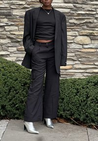 Zwarte blazer over een zwarte crop top, gecombineerd met zwarte wijde cargo broeken en metallic zilveren enkellaarsjes. Achtergrond van een gecementeerde stenen muur.