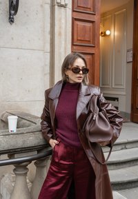 Palton oversized din piele maro, pulover roșu închis cu fante, pantaloni de satin burgund, și o geantă maro. Ceașcă albă de cafea pe balustrada de piatră.