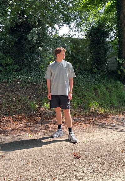 Camiseta gris de gran tamaño, pantalones cortos negros, zapatos deportivos grises, de pie en un sendero de tierra rodeado de vegetación. La luz del sol filtra a través de los árboles.