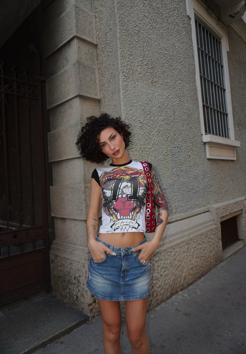 Femme aux cheveux bouclés debout contre un mur de pierre texturé, portant un crop top graphique, une jupe en denim et un sac épaule rouge, les mains dans les poches.
