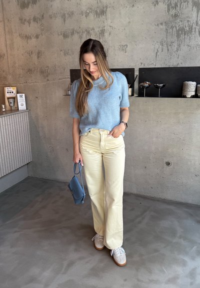 Suéter de punto azul claro, pantalones de pierna ancha color crema, zapatillas grises con suelas marrones y un pequeño bolso azul. Fondo de concreto con una estantería.