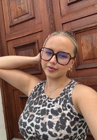 Top na ramiączkach w wzór panterki z prążkowanej tkaniny, w zestawieniu z dużymi okularami w tortoishell, złotą biżuterią, na tle drewnianych drzwi.