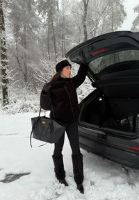 Cacală de blană sintetică neagră cu nasturi mari, colanți negri și cizme de blană până la genunchi. Ținând o geantă texturată neagră lângă o mașină într-o pădure acoperită de zăpadă.