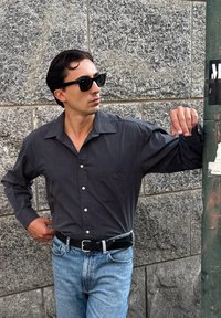Camisa oscida de botones y manga larga, combinada con jeans azul claro y un cinturón negro. Lleva gafas de sol negras y está apoyado contra una pared.