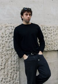 Maglietta nera a maniche lunghe e jeans scuri; in piedi contro un muro di pietra strutturato con un'area chiara e ruvida, mani nelle tasche.