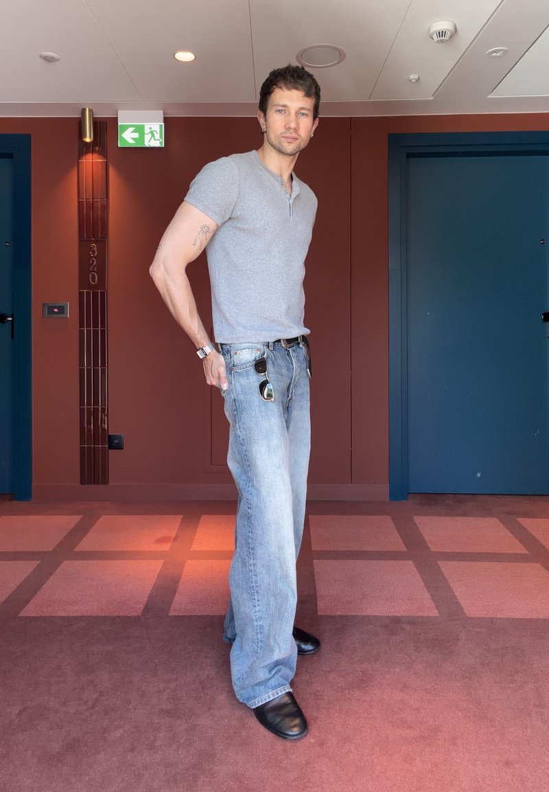 Homme en chemise grise et jean bleu ample debout dans un couloir aux murs rouges et portes bleues, tenant des lunettes de soleil accrochées à son jean.