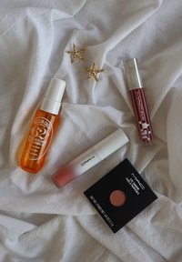Bruma para el cabello en una botella naranja, producto labial blanco, sombra de ojos en un estuche negro y dos pendientes dorados en forma de estrella sobre una tela blanca.