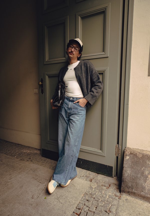 Person læner sig op ad en grå dør iført briller, en kasket, hvid skjorte, grå cardigan, vide blå jeans og hvide sneakers, holder en sort taske.