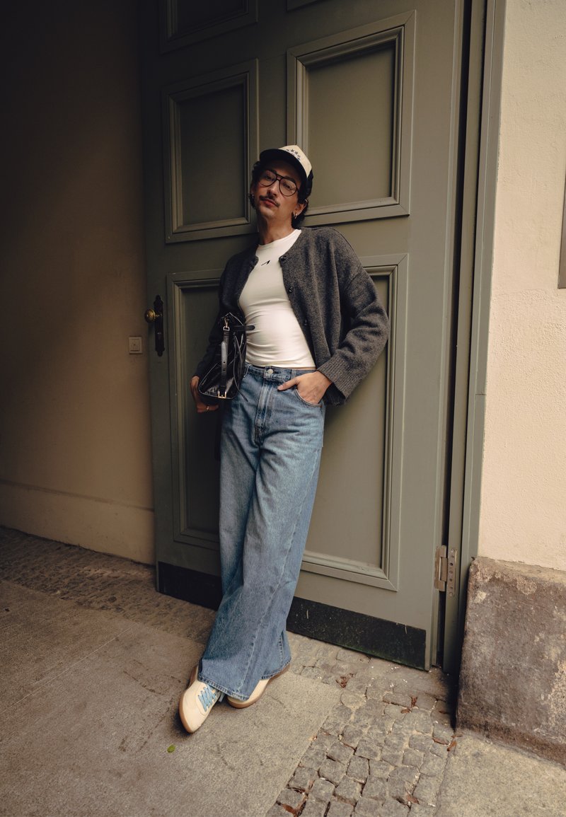 Personne appuyée contre une porte grise, portant des lunettes, une casquette, une chemise blanche, un cardigan gris, un jean bleu large et des baskets blanches, tenant un sac noir.