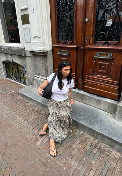 Bolso de tote negro, camiseta blanca y falda larga de estampado de leopardo. Sentada en un escalón de piedra frente a una puerta de madera, rodeada de adoquines.