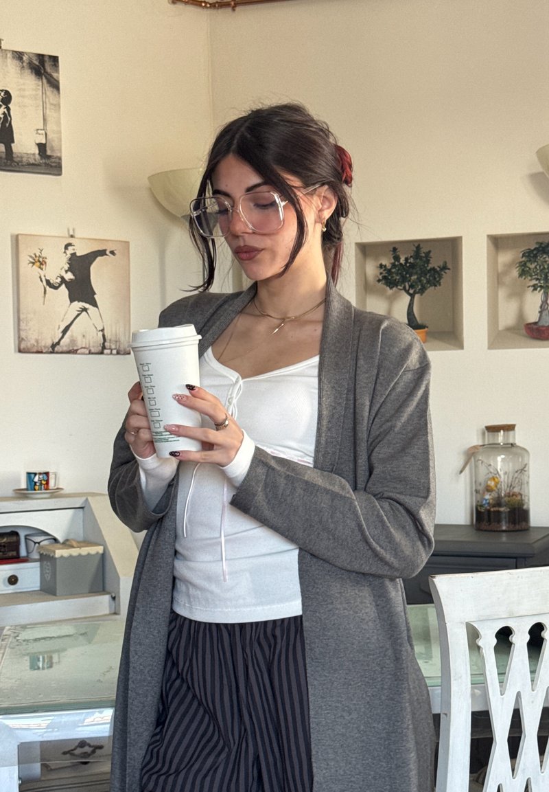 Cardigan gris sur un haut blanc, tenant une tasse de café. Cheveux longs et foncés avec des accents rouges, grandes lunettes transparentes, pantalon à rayures noir et blanc.