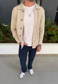 Bežs adīts uzvalks ar pogām virs baltā t-kreklu, apvienots ar tumši ziliem džinsiem un pelēkbaltiem sporta apaviem. Iekļautas sudraba rotaslietas un dabīgs fons.