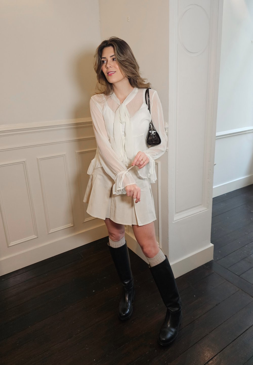 Ljus, genomskinlig vit blus med en rosett, lager av rynkig nederkant, kombinerad med en beige utsvängd kjol, knähöga svarta stövlar och en svart handväska.