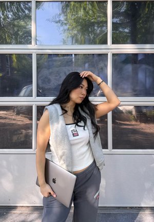 Chaleco sin mangas gris sobre una camiseta blanca con parche de "USA", combinado con pantalones de chándal grises, sosteniendo una computadora portátil plateada contra un fondo de cristal.