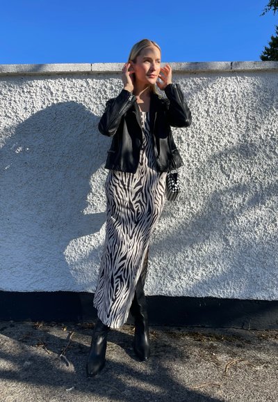 Chaqueta de cuero negra sobre un vestido largo de maxi estampado de cebra en beige y negro, combinado con botas negras de tacón alto y un bolso estampado.