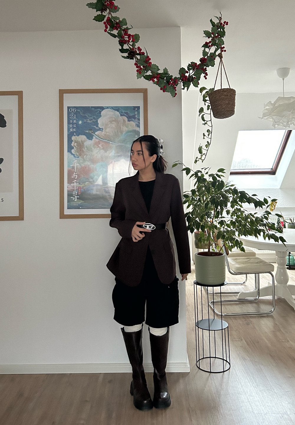 Blazer marron sur un haut noir, associé à un short noir, des bottes marron hauteur genou et une boucle de ceinture argentée en accent. Cadre intérieur avec de la verdure.