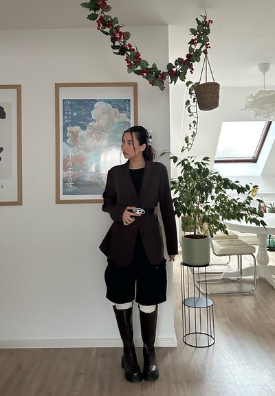 Blazer marrón sobre una camiseta negra, combinada con pantalones cortos negros, botas color chocolate hasta la rodilla y un hebilla de cinturón plateada como detalle. Ambiente interior con plantas.