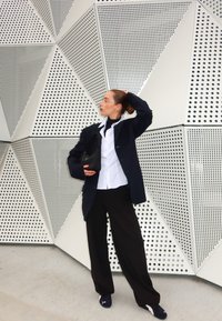 Svart oversized blazer över en vit skjorta och svarta byxor, håller en svart handväska, mot en geometrisk perforerad väggbakgrund.