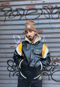 Μαύρο δερμάτινο μπουφάν τύπου bomber με κίτρινες και λευκές λεπτομέρειες, με κεντημένο λογότυπο. Συνδυάζεται με καφέ πλεκτό σκουφί και μπεζ φούτερ.