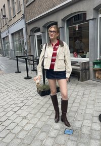 Beige avkortet jakke med brun krage, rødstripet polo-shirt, denim mini skjørt, brun støvel til kneet, holder en kaffekopp, på brosteinsgate.