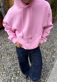 Rosa genser med hette og frontlomme, med snorer og løs passform, kombinert med vide mørkeblå jeans.